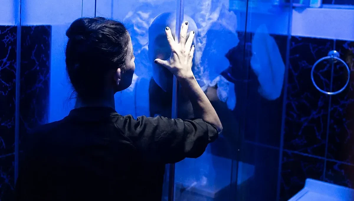 Pessoa vista de costas toca com a mão um painel de vidro em um ambiente iluminado em tons de azul; sua imagem aparece refletida no vidro.