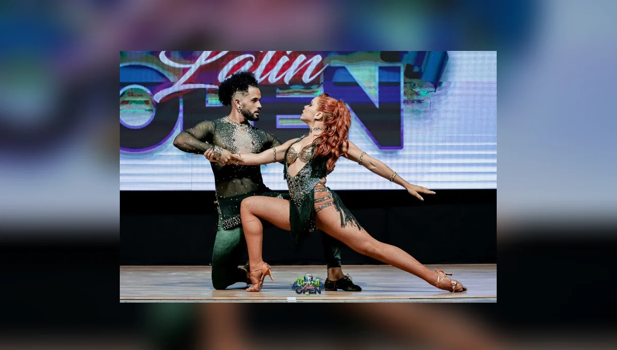 Um homem e uma mulher dançam em pose coreografada no palco, diante de um painel iluminado.