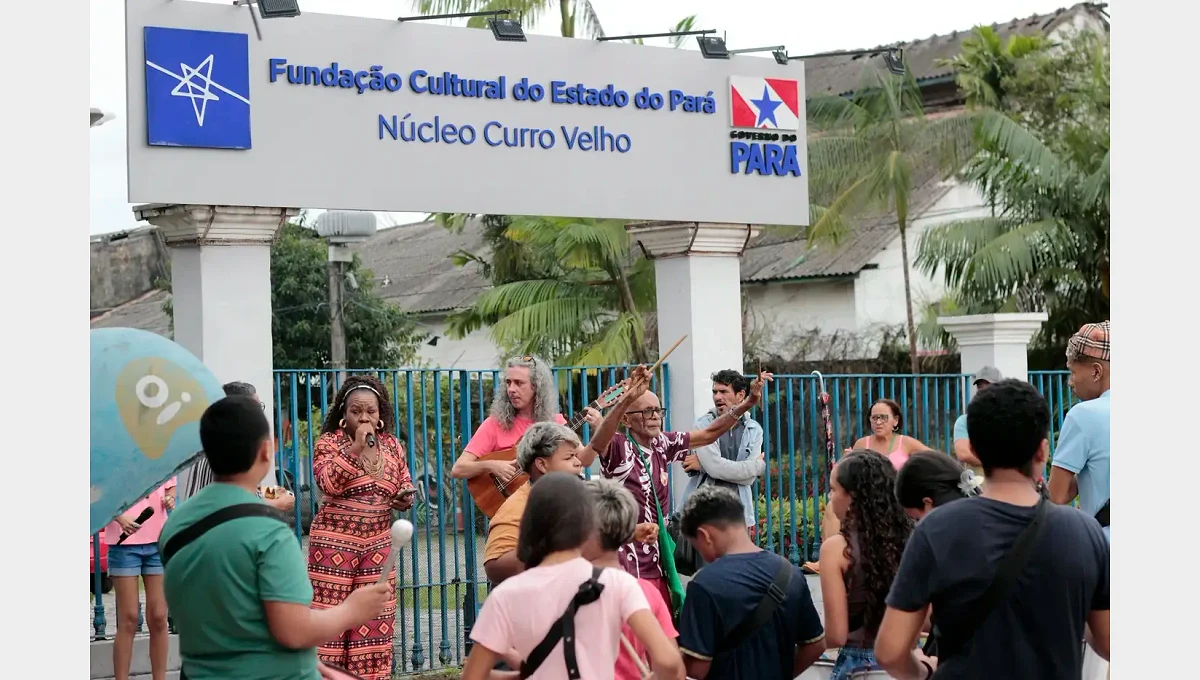 Pessoas reunidas diante do portão do Núcleo Curro Velho, sob a placa da instituição, com instrumentos musicais.