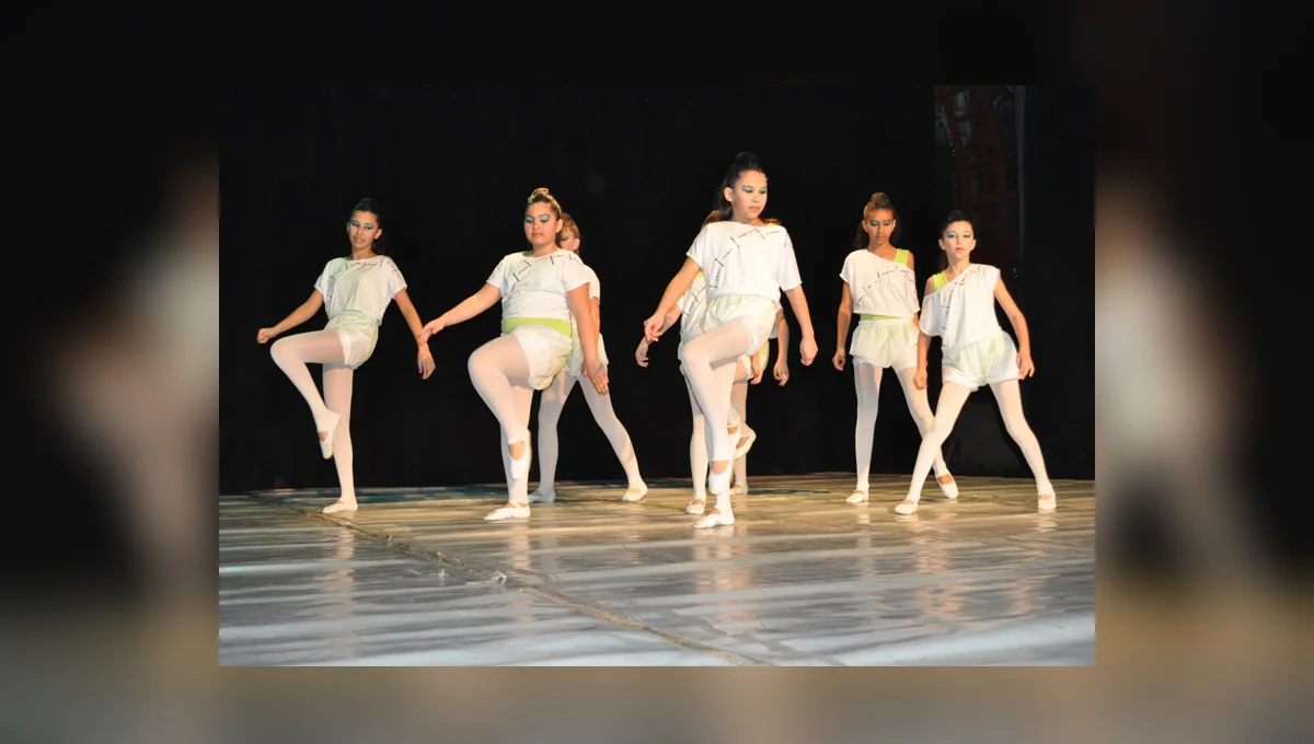 Seis bailarinas em figurinos brancos se apresentam em formação sobre um palco iluminado, diante de cortinas escuras.
