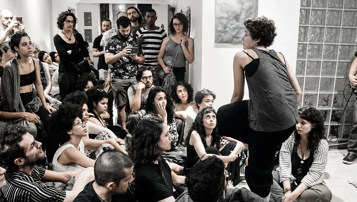Em uma sala com obras nas paredes, uma mulher está de pé diante de um grupo de pessoas sentadas no chão.