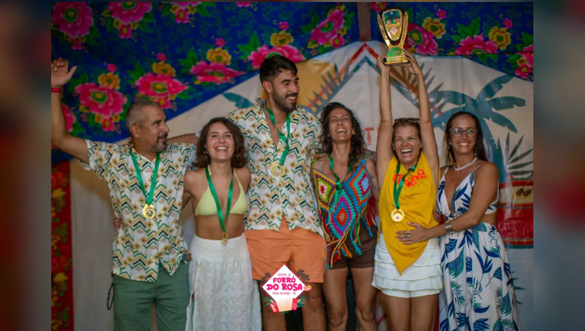 Grupo de pessoas posa junto a um troféu; uma delas o ergue. Algumas usam medalhas, diante de um painel colorido com a marca Forró do Rosa.
