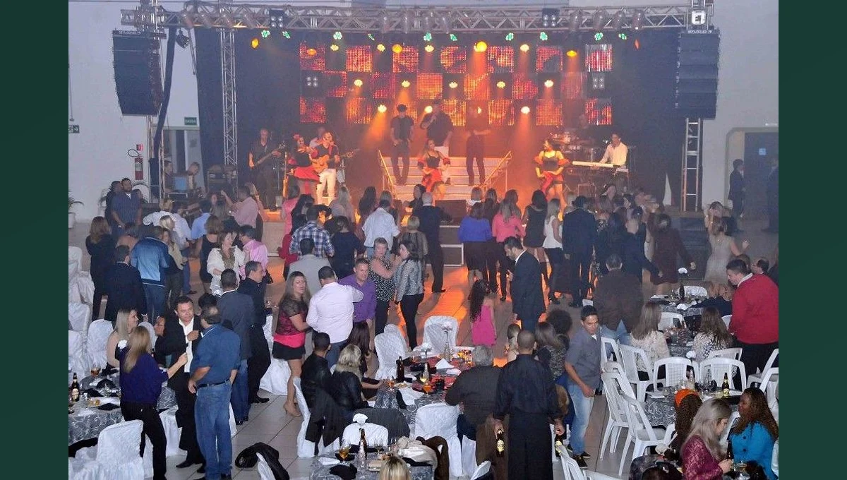 Público dança entre mesas diante de uma banda em um salão iluminado.
