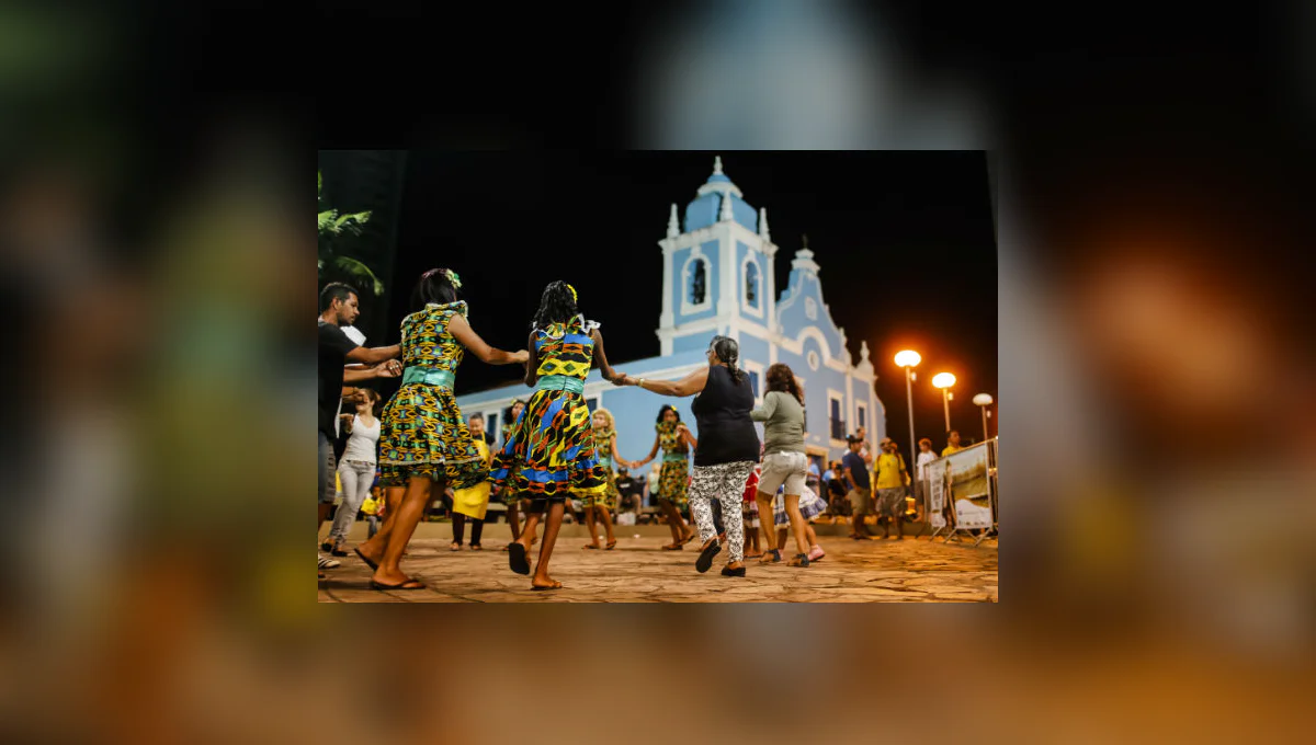 Pessoas de mãos dadas dançam em roda diante de uma igreja iluminada à noite.