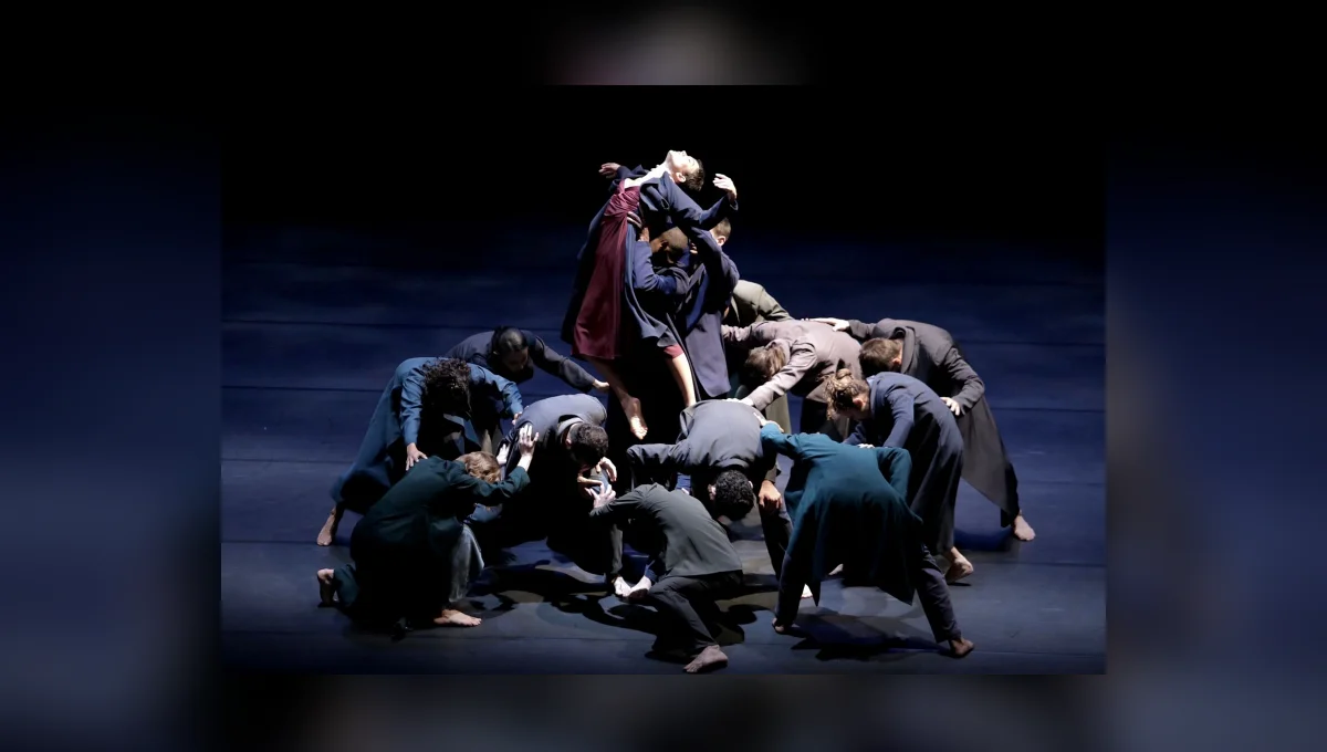 Grupo de bailarinos forma uma composição no palco, com alguns dançarinos agachados ao redor de outros que se erguem no centro.